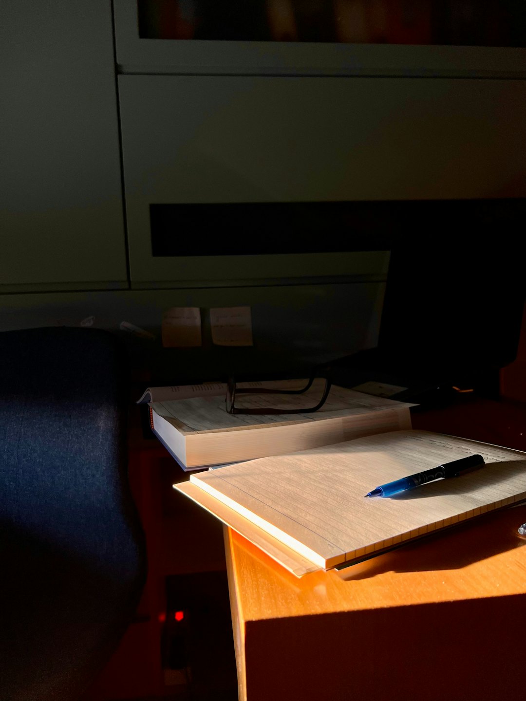 Wooden desk with open book, glasses, notebook, and pen in sunlight.
